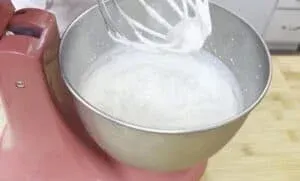 Mixer whipping cream in a bowl, preparing ingredients for baking on a wooden countertop.